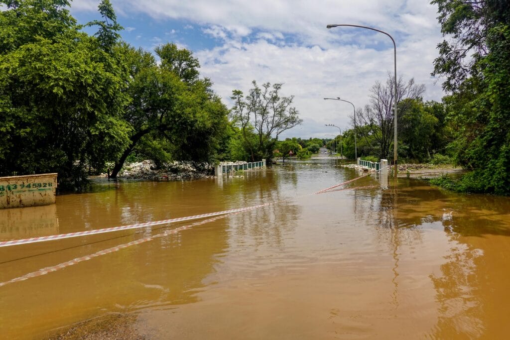 A flooded road with a submerged bridge, surrounded by trees, under a cloudy sky.